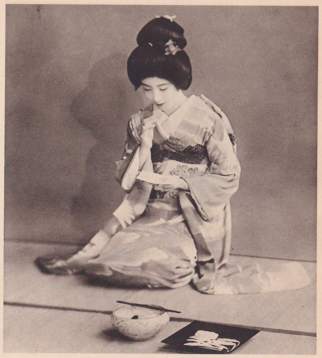 Photograph of a woman with her hair in a high bun with ornaments, in a kimono with pale cloud shapes, kneeling and facing the camera with her head bowed, one hand raised near her mouth and a folded sheet of paper in the other. On the mat before her stand a pale bowl with a thin stick laid across its rim and a dark square tray with pale objects on it.