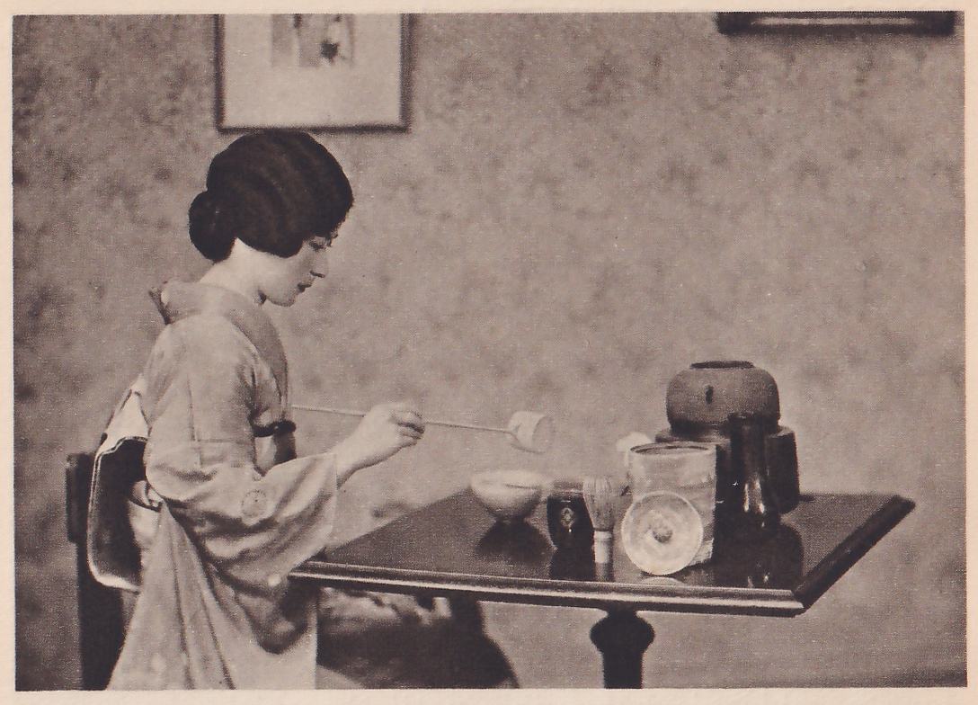 Photograph of a woman with her hair in a bun, in a pale kimono, seated on a chair in profile facing right at a small dark square table, one hand holding a long thin-handled ladle out over a pale bowl; on the table are the bowl, a whisk, a small dark pot, a pale lidded jar and a large round dark pot. Floral wallpaper and a framed picture are behind her.