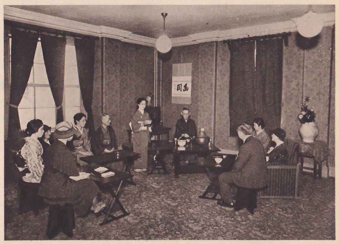 Photograph of a Western-style room with curtained windows, a patterned carpet, ceiling lamps and a hanging scroll of writing on the far wall. People sit on stools and chairs at low folding tables, some in Western dress and some in kimono; at the center a woman in a kimono stands with her hands together at her chest, and a man in dark clothes sits behind a low table with a kettle and bowls. A vase of flowers stands on a stand at the right.