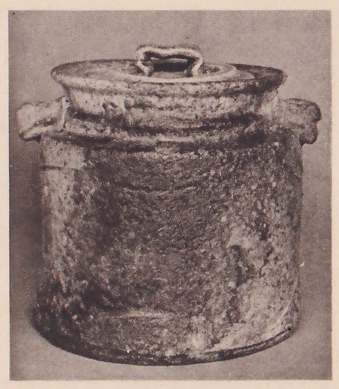 Photograph of a squat rough-glazed jar with speckled brown sides, a flat lid topped by a loop-shaped knob and two small lugs at the sides, on a plain gray ground.