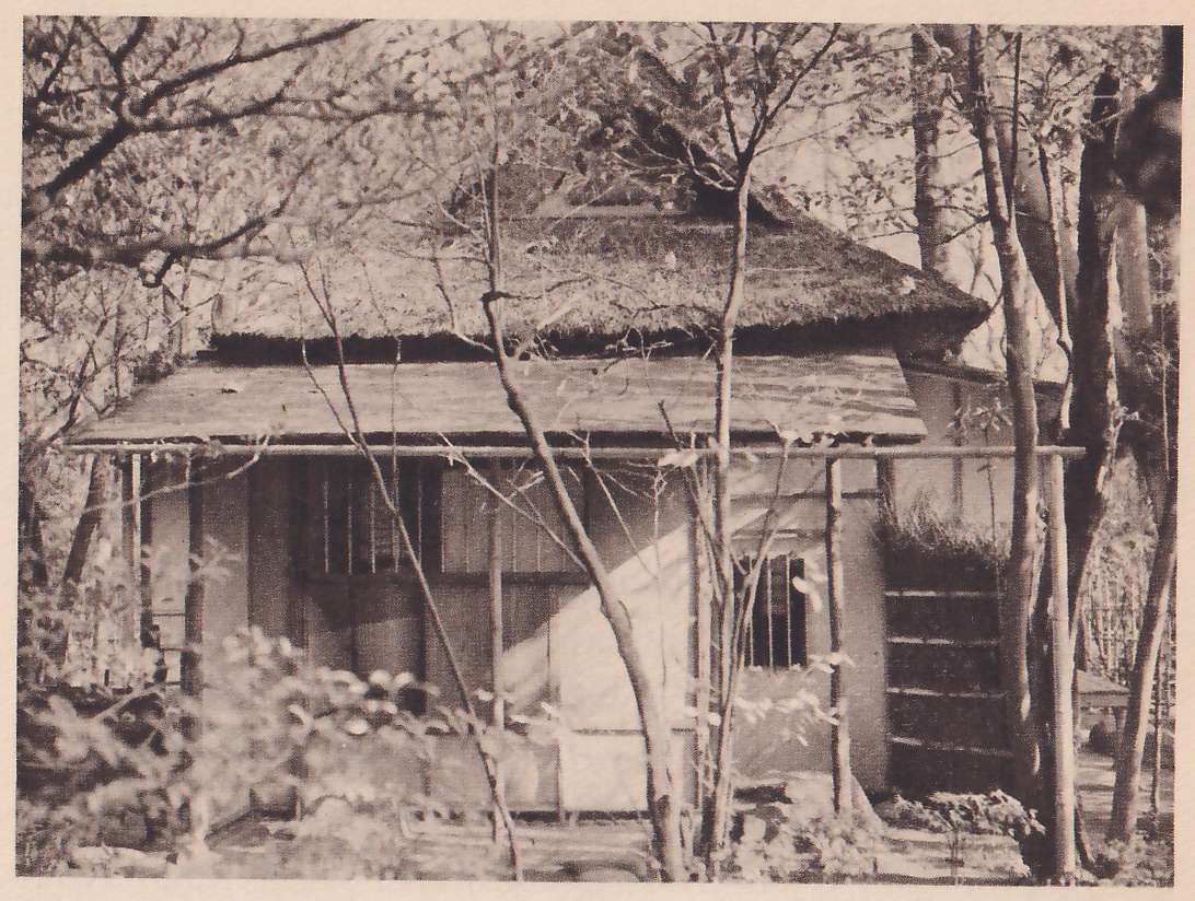 Photograph of a small house with a deep thatched roof and a lower sloping roof over a front veranda, seen through bare branches and tree trunks; the front has a pale board wall and two barred windows, and at the right a screen of horizontal slats.