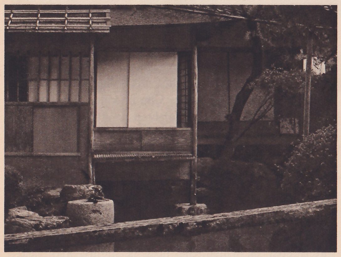 Photograph of the side of a wooden house raised on posts over a pond: a pair of pale sliding paper doors above a plank rail, a latticed window at the left, and dark space beneath. In the foreground the stone edge of the pond, at the left a round stone basin among rocks, and at the right a clipped shrub and a tree trunk.