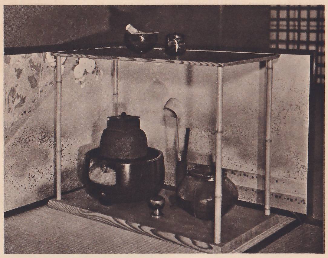 Photograph of a two-shelf stand on four slender jointed poles in the corner of a mat room before a screen painted with flowers: on the upper shelf a dark bowl with something white laid across it and a small dark jar; on the lower shelf a large dark iron kettle on a round dark brazier, a bamboo ladle standing beside it, a dark jar and a small metal object.