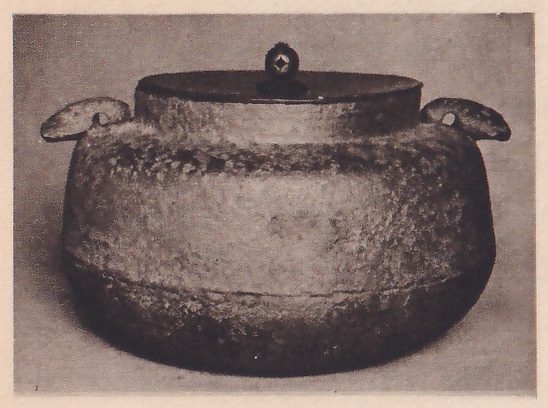 Photograph of a squat iron kettle with a rough surface, a flat lid topped by a small ring-shaped knob, a band around its lower body and two small ear-shaped handles at the shoulders, on a plain gray ground.