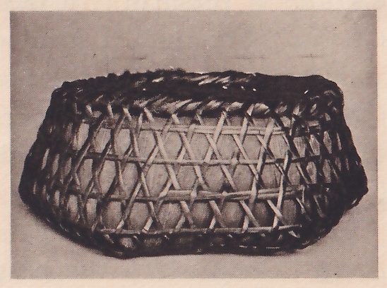 Photograph of a low shallow basket woven of split bamboo strips in a lattice of crossing diagonals with a dark rim, wider at the top than at the base, on a plain gray ground.