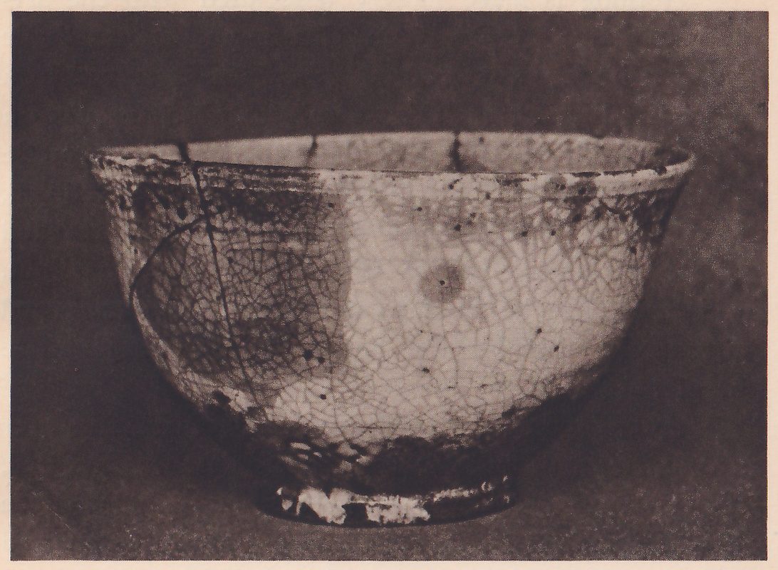 Photograph of a tea bowl seen from the side on a dark ground: a broad bowl on a small foot ring, its pale crackled glaze patched with darker areas, a thin dark line running down the left side and a small round brown spot at the center.
