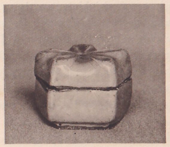 Photograph of a small squat lidded container on a plain gray ground: pale flat sides, a darker rim between lid and base, and a lid that slopes up to a small knob at the top.