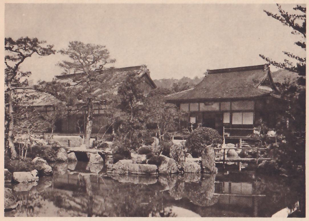Photograph across a still pond of a low wooden building with a wide sweeping roof at the right and a second roof among pine trees at the left; rocks line the pond's edge with a small plank bridge between them, and the buildings and trees are reflected in the water.