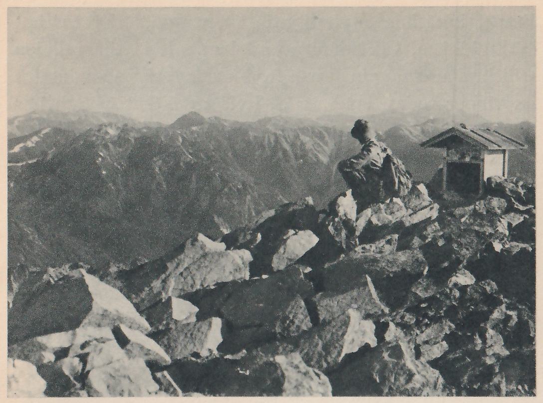 Photograph of a person seen from behind, sitting on a heap of broken rocks on a mountain top and looking out over ranges of peaks; a very small roofed shrine stands on the rocks to the right of the person.