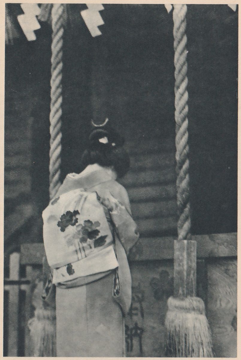 Photograph from behind of a person in a pale kimono with a wide flower-patterned sash and hair put up with a small pale ornament, standing before the dark wooden front of a shrine and a wooden offertory box, between two thick twisted ropes that end in tassels; zigzag strips of white paper hang at the top.