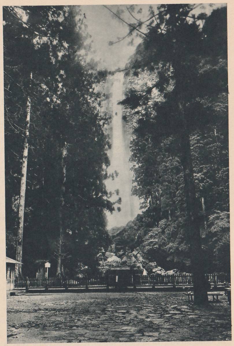 Photograph of a very tall, thin waterfall dropping from the top of the picture between dark trees, seen from a stone-paved open area; a low fence and small roofed buildings stand across the far side of the open area, below the trees.