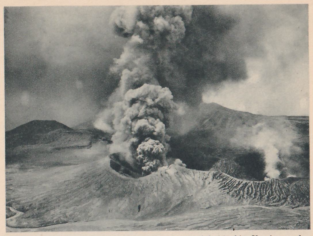 Photograph of a volcano: a thick column of dark smoke and ash rises from a crater in the foreground, with a wide crater rim around it, other cones behind, and plumes of steam at the right.