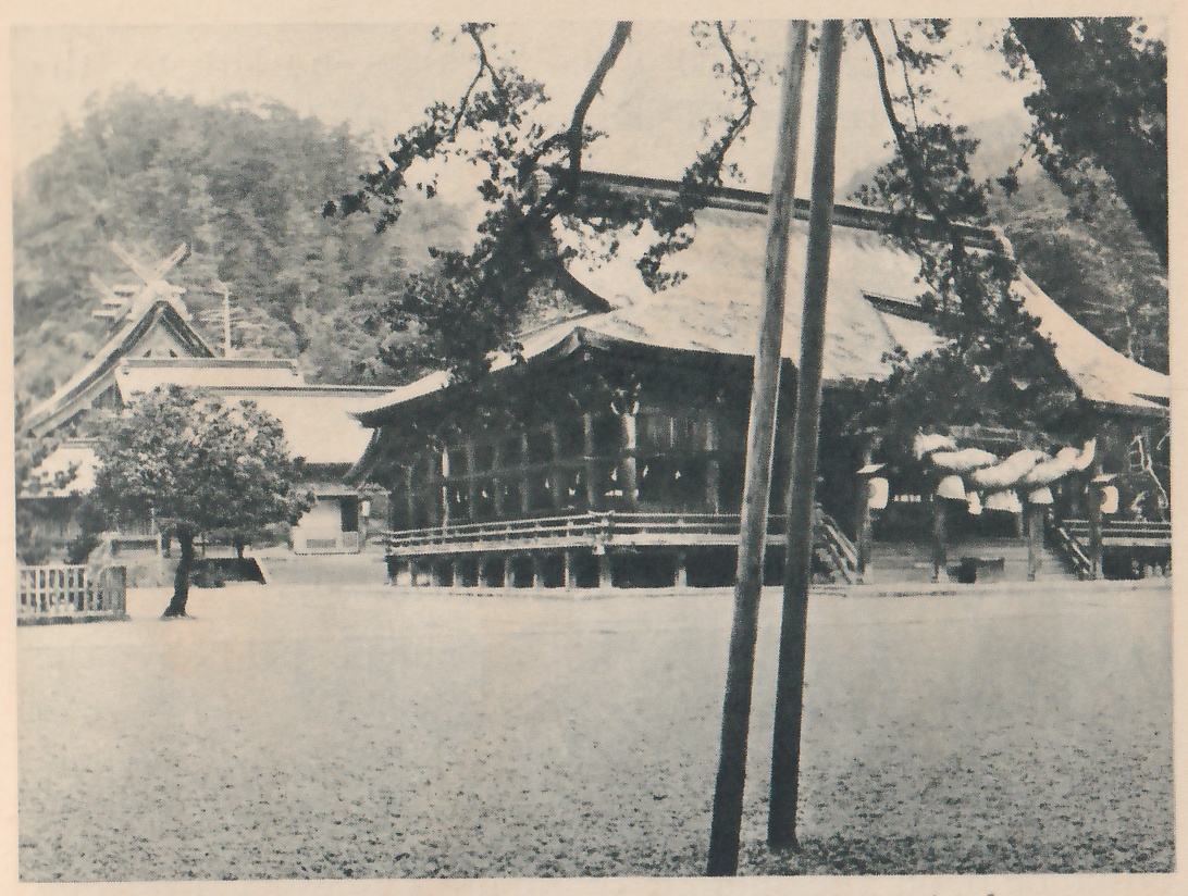 Photograph of a wide wooden hall with a heavy sweeping roof and a railed porch, with pale rounded shapes hanging under the eaves at the right; behind it at the left stands a building with crossed beams at the roof ridge, and two tall thin poles lean together in the foreground over an open area of gravel.