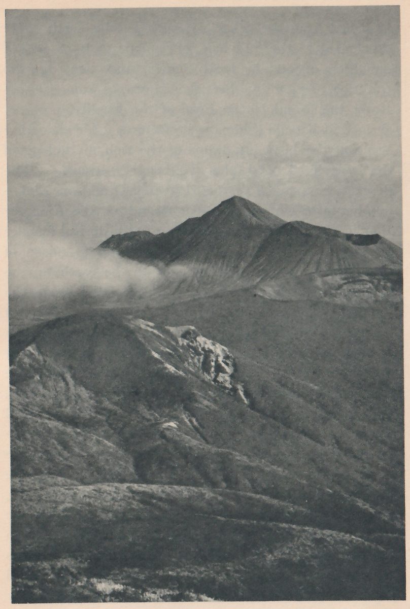Photograph looking across dark slopes to a mountain with a pointed peak and a lower cone with a crater beside it, with a band of cloud lying at the left of them.