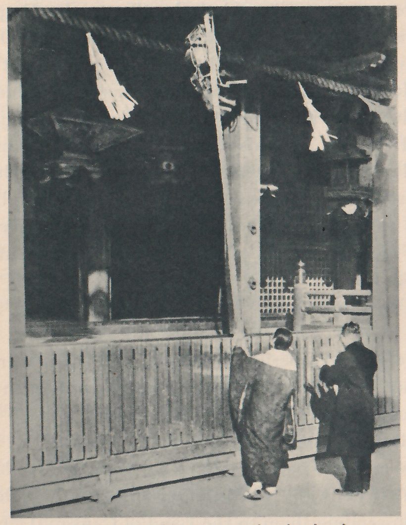 Photograph of two people seen from behind, standing close together before the dark open front of a shrine behind a low slatted fence; the one at the left wears a long patterned robe with a pale collar, the one at the right a long dark coat; a thick rope with zigzag strips of white paper hangs across the top, and a long straight band slants down from an ornament hanging at the top to the raised hand of the person at the left.