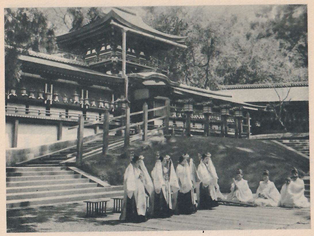 Photograph of a group of people in long white over-robes and long dark skirts, with ornaments in their hair, standing in a line at the left, and several more in white sitting on a wooden platform at the right, in front of a stairway and a roofed hall with rows of lanterns under its eaves.