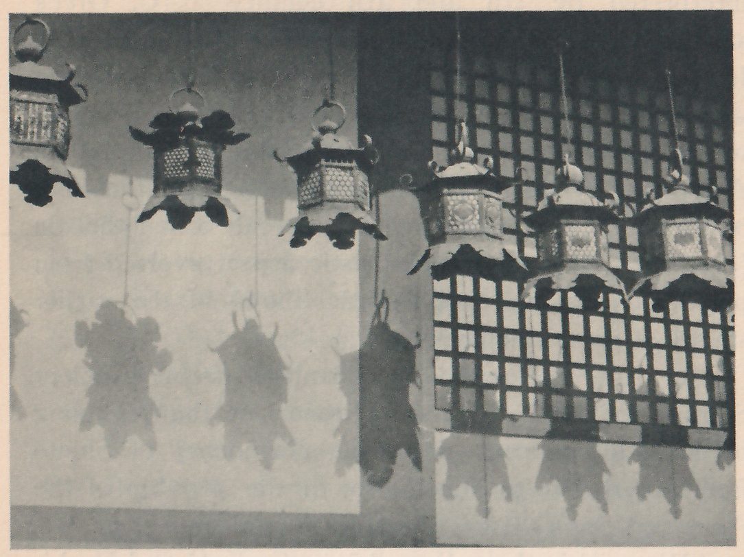 Photograph of a row of metal lanterns hanging from above, each with a flared roof and pierced sides, with their dark shadows on the pale wall below; a wooden lattice panel fills the right side.