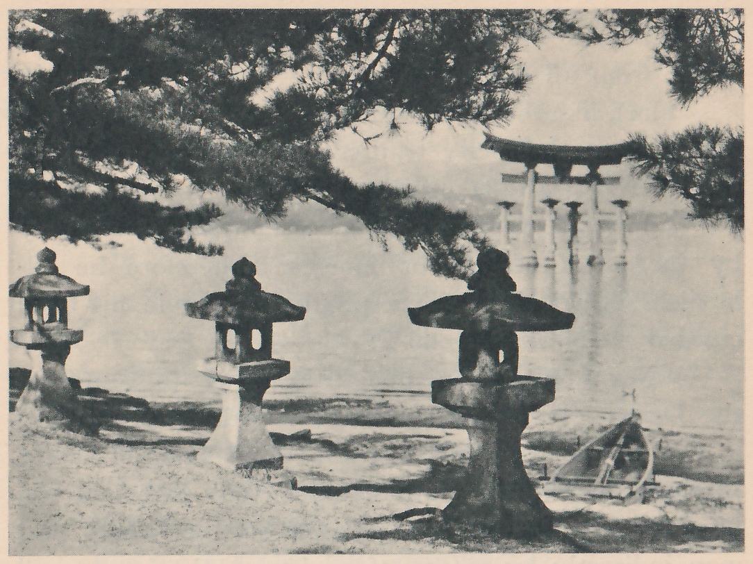 Photograph through pine branches at the upper left of a shore: stone lanterns stand on the sand in the foreground, a torii gate stands out in the water at the right of center, and a small boat lies on the sand at the lower right.