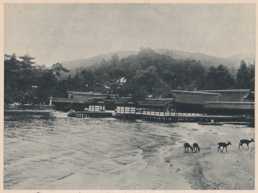Photograph across a bay of a long, low shrine building standing at the water’s edge below wooded hills, with the top of a pagoda showing at the upper left and a few deer on the sand at the lower right.