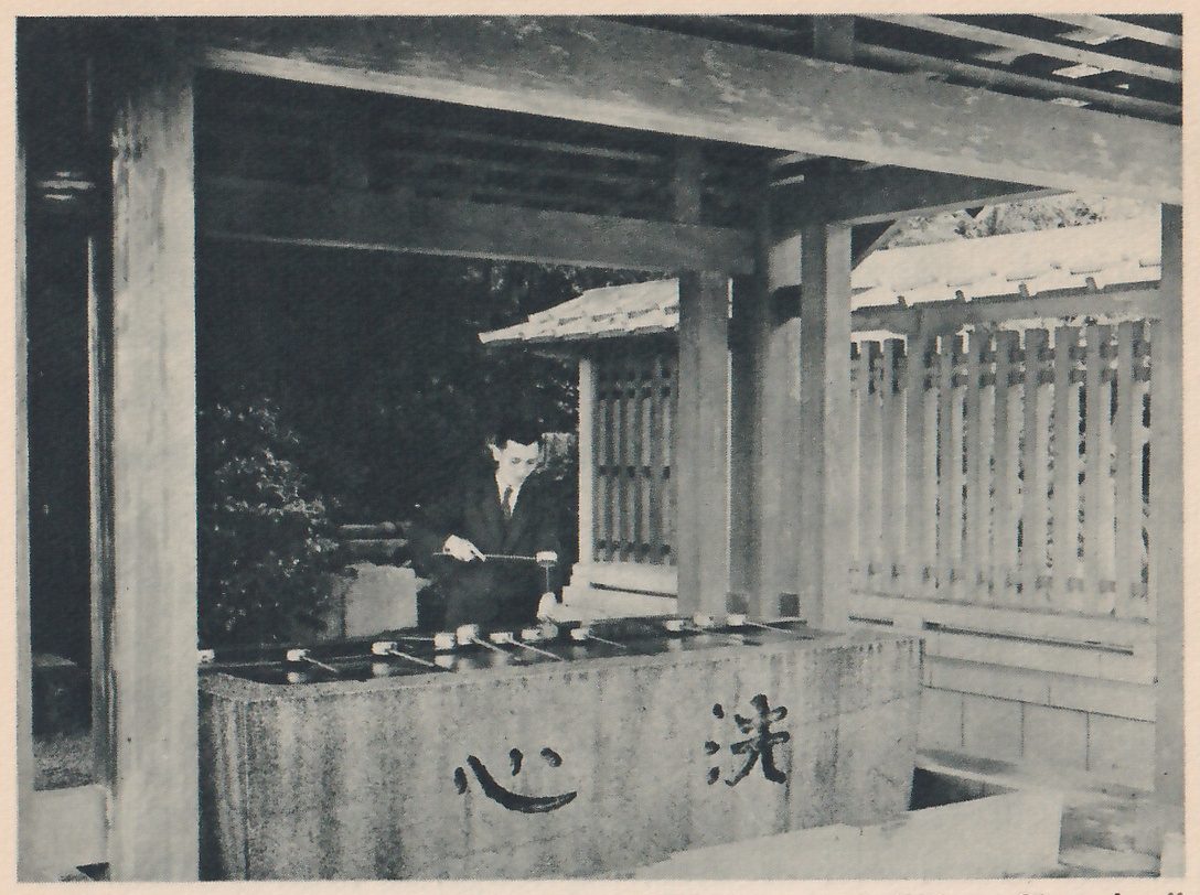 Photograph of an open washing pavilion with thick wooden pillars and a large stone basin; several long-handled ladles lie across the basin, a man in a dark suit and tie behind it holds a ladle out over the basin, and two large characters carved on the front of the basin read senshin, heart-cleansing.