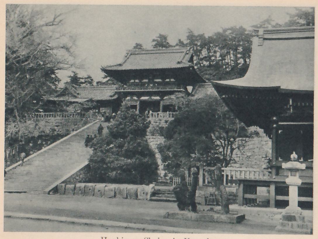 Photograph of a broad stone stairway rising from the lower left to a two-story gate with a curved tiled roof at the center, other buildings behind it, and the near corner of a large roof at the right with a stone lantern beside it.