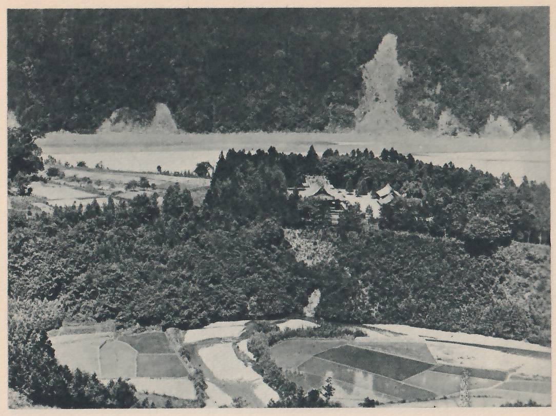 Photograph taken from a height of a cluster of low roofed buildings in a clearing among dense dark trees on a hillside, with small fields in the foreground and a rocky slope in the background.