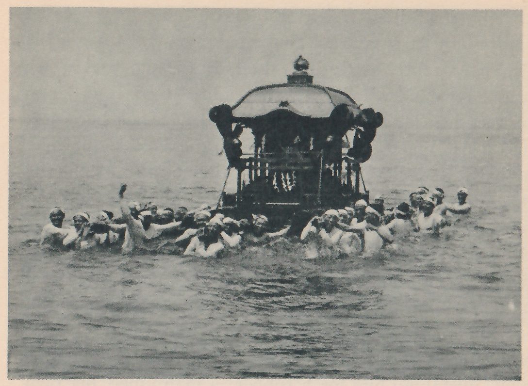 Photograph of a crowd of men standing in water, many with white cloths on their heads, with a portable shrine with a domed roof, a small finial on top and black round ornaments held above them; one man’s arm is raised at the left.