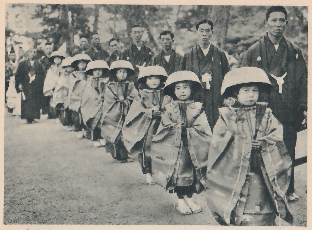 Photograph of a line of children in stiff, long patterned robes with cords at the chest and wide shallow round hats, standing one behind another in a row that runs from the lower right back toward the upper left; the nearest faces the camera. Behind and beside them stand adults in dark kimono, several with a pale badge on the chest.