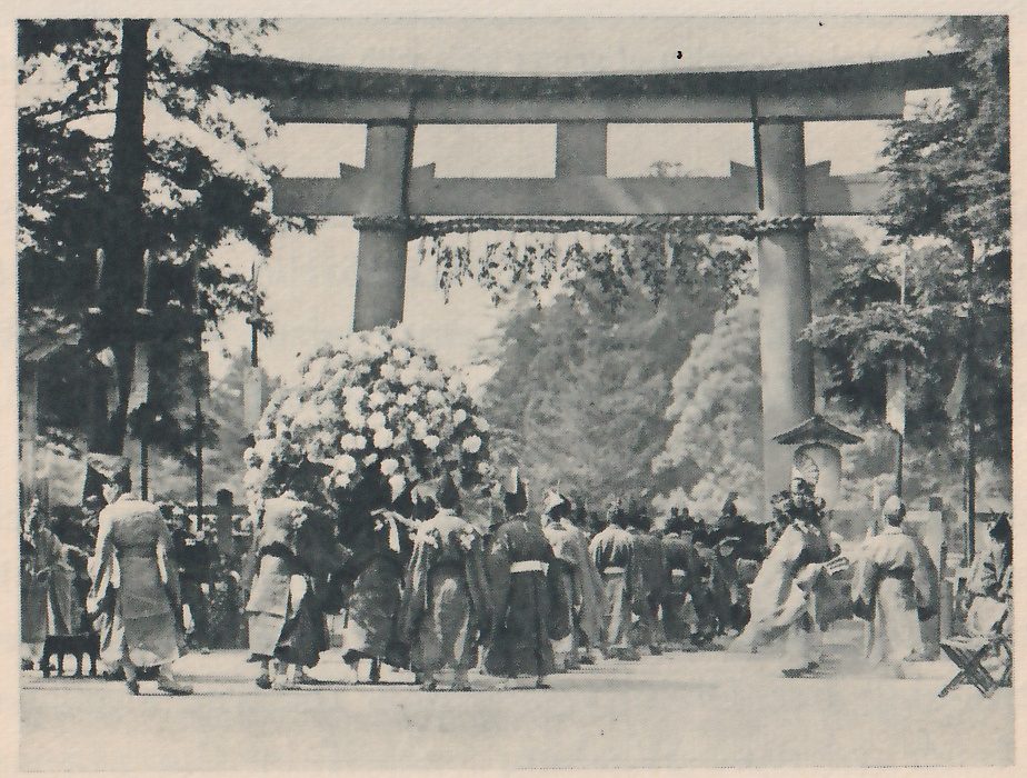 Photograph of a large torii gate with a heavy double top beam standing over a crowd of people in long robes of several kinds; a large rounded mass of pale leaves or blossoms rises above the crowd at the left of center.