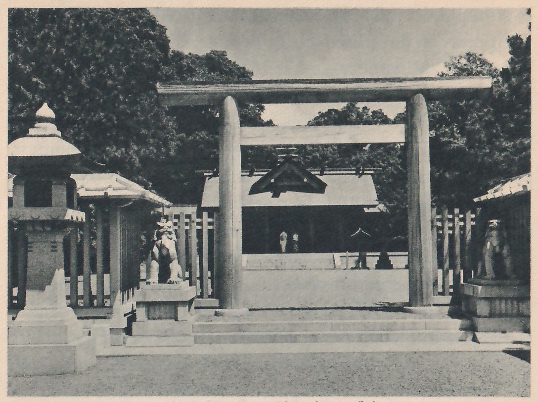 Photograph of a wooden torii with a top beam and a lower crossbeam in front of a long low hall with a gabled front and low steps; a stone lantern stands at the left, and a stone guardian figure sits on a pedestal on each side of the path.
