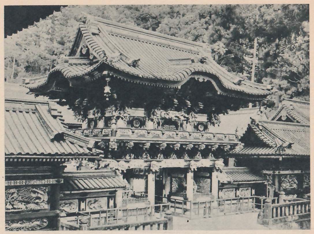 Photograph of an ornate gate with a curved tiled roof above richly carved and decorated upper walls, with other tiled roofs, carved panels and a railing around it.
