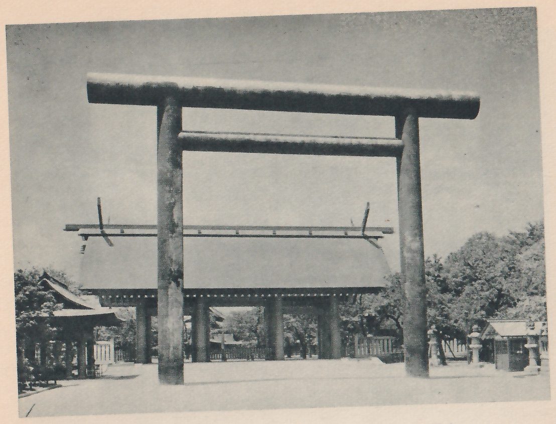 Photograph of a large torii with thick round pillars, a heavy top beam and a lower crossbeam, standing in front of a long low gate whose wide roof has crossed beams sticking up at the ridge ends; a small roofed building shows at the left, stone lanterns and a small roofed hut at the right, and trees around.