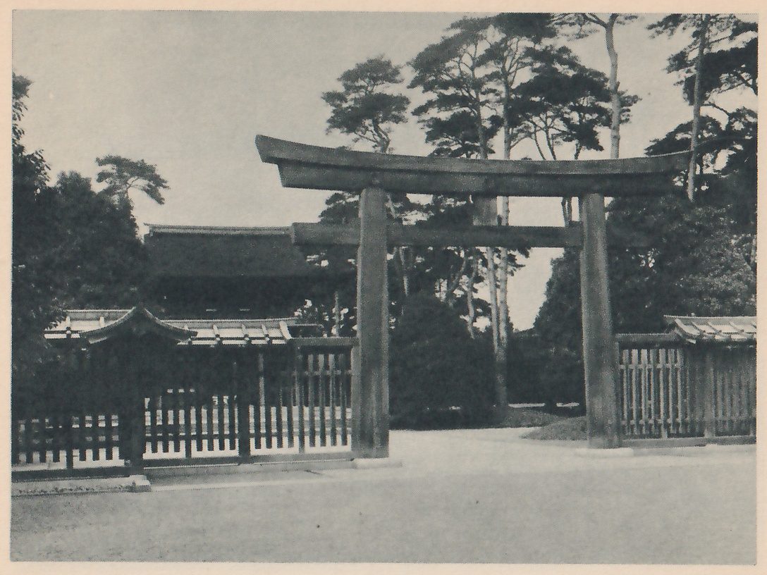 Photograph of a dark wooden torii with a curved top beam at the right, standing before a low wooden fence and small roofed gate buildings, with pine trees behind and the large roof of a hall showing in the middle distance.
