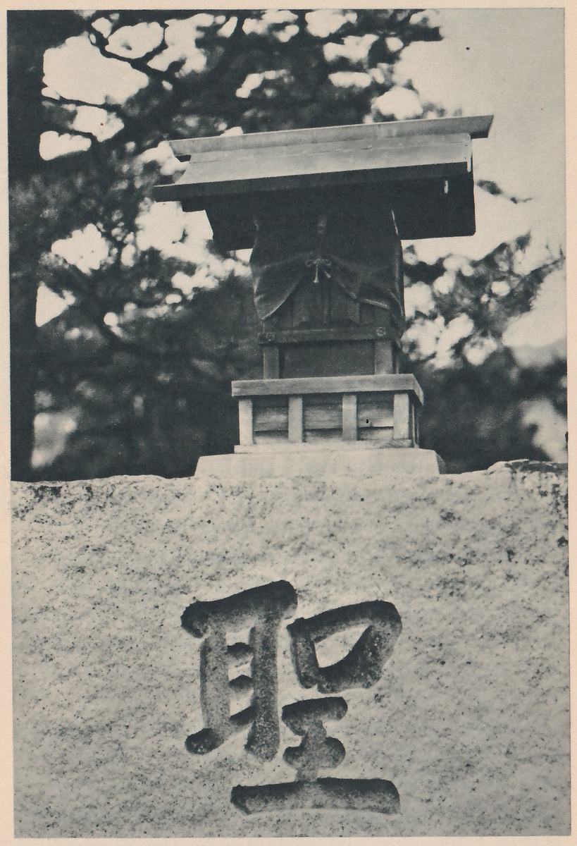 Photograph of a small wooden shrine with a roof, set on a low platform on top of a block of stone; one large character carved into the front of the stone reads “holy”, and pine branches are out of focus behind.
