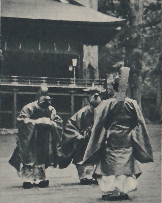 Photograph of at least three men in wide-sleeved robes over pale trousers in a courtyard before a wooden hall with a railed veranda; the two at the left, each in a small dark cap, bend forward facing the camera, and the man at the right, seen from behind, wears a tall stiff hat.